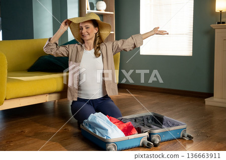 Happy woman wearing a straw hat and smiling broadly, sitting on the floor in her living room, packing clothes into an open suitcase, anticipating an exciting journey 136663911