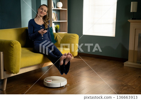 Woman relaxing on a sofa with bare feet up, scrolling a smartphone while a robot vacuum quietly cleans the wooden floor, illustrating effortless smart home comfort and convenience Woman relaxing on a sofa with bare feet up, scrolling a smartphone while a robot vacuum quietly cleans the wooden floor, illustrating effortless smart home comfort and convenience 136663913