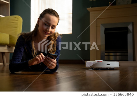 Young woman lying on the living room floor, smiling and using a smartphone, while a robotic vacuum cleaner works nearby, symbolizing modern convenience, smart home technology, and effortless chores Young woman lying on the living room floor, smiling and using a smartphone, while a robotic vacuum cleaner works nearby, symbolizing modern convenience, smart home technology, and effortless chores 136663940