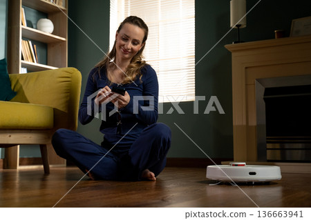 Woman sitting on floor, relaxing while controlling a smart robot vacuum cleaner in living room using a mobile phone application for automated home cleaning Woman sitting on floor, relaxing while controlling a smart robot vacuum cleaner in living room using a mobile phone application for automated home cleaning 136663941