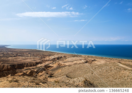 Scenic view of Aksai Canyon with its rugged landscape bordering the calm blue water of Issyk Kul lake under a clear sky for travel brochure. Kyrgyzstan. 136665248