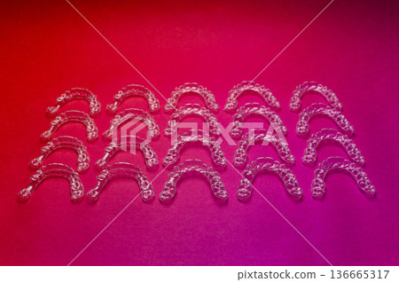 Clear dental aligners arranged in rows on a vibrant pink background, showcasing their transparent design and orthodontic purpose for teeth alignment 136665317