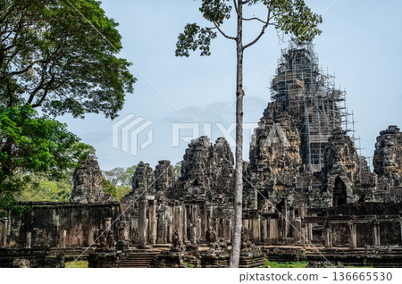 Restoration work on ancient Bayon temple face towers with metal scaffolding and tall tree in foreground under blue sky background 136665530