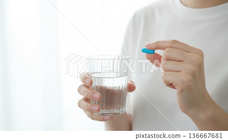 Woman in white shirt holding a blue medicine capsule and a clear glass of water in bright light. 136667681