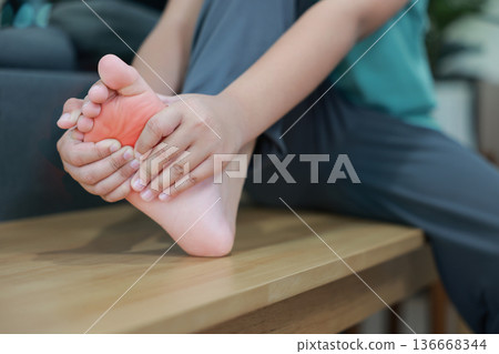 Close up of Asian woman holding her foot with red highlight on painful area, suffering from plantar fasciitis, foot pain, numbness, peripheral neuropathy, muscle strain or inflammation at home. 136668344