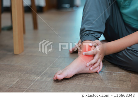 Close up of child's hands holding ankle with red highlight on painful area, Asian boy suffering from ankle sprain, foot injury, ligament strain or bone inflammation sitting on floor. 136668348