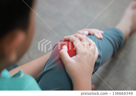 Asian boy showing painful face expression holding red inflamed knee from joint injury or accident. Pediatric healthcare concept of childhood bone problems, growth pain, and orthopedic treatment. 136668349