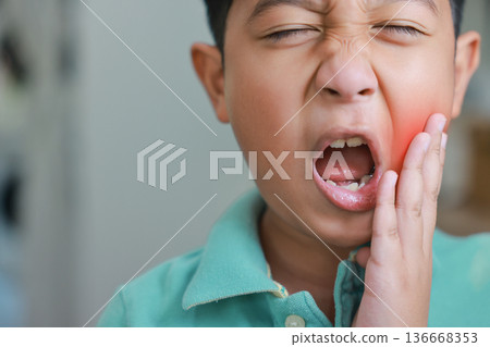 Asian boy suffering from severe toothache with red highlight on cheek, child having dental problem, tooth decay, gum inflammation, sensitive teeth or wisdom tooth pain and crying at home. 136668353