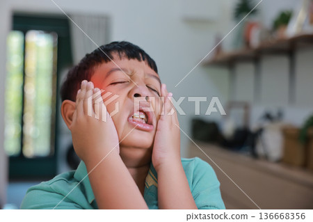 Asian boy suffering from severe headache or migraine with red highlight on forehead, child having fever, dizziness, brain tumor symptoms or stress while touching head at home. 136668356