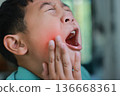 Asian boy suffering from severe toothache with red highlight on cheek, child having dental problem, tooth decay, gum inflammation, sensitive teeth or wisdom tooth pain at home. 136668361