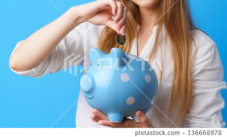 Woman puts a coin into a blue piggy bank with white dots on a blue background Woman puts a coin into a blue piggy bank with white dots on a blue background 136668978