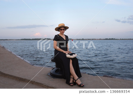 Fashionable woman sits on mooring post by calm water. 136669354