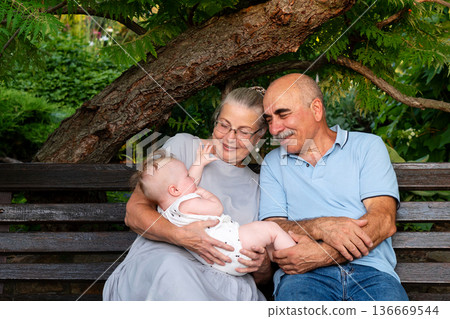 Elderly couple joyfully holding baby on garden bench Elderly couple joyfully holding baby on garden bench 136669544