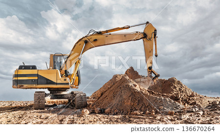 A large excavator digs into a mound of dirt at a work site on a cloudy day in the afternoon. The machine is in action, moving earth 136670264