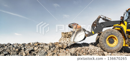 At a construction site, a yellow loader or bulldozer uses its bucket to lift and move large rocks. The sky is clear, the terrain is rocky, and there are no trees At a construction site, a yellow loader or bulldozer uses its bucket to lift and move large rocks. The sky is clear, the terrain is rocky, and there are no trees 136670265