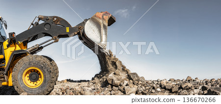 Construction equipment, such as a loader or bulldozer, lifts rocks and dumps them onto a pile at a construction site. Earthworks are used to form and level the construction site Construction equipment, such as a loader or bulldozer, lifts rocks and dumps them onto a pile at a construction site. Earthworks are used to form and level the construction site 136670266
