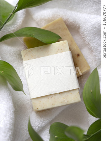 Mockup of beige solid soap bar with unbranded paper label near green leaves on white bath towels 136670777