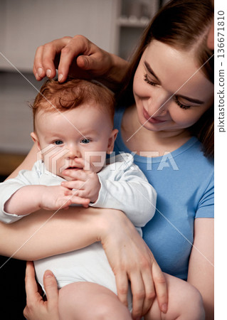 Mother holds her baby in her arms while smiling in a room with warm lighting and cozy atmosphere during an afternoon family moment 136671810
