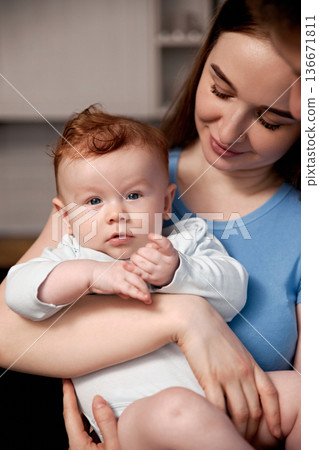Mother holds baby in her arms while smiling in a cozy indoor setting during daytime 136671811