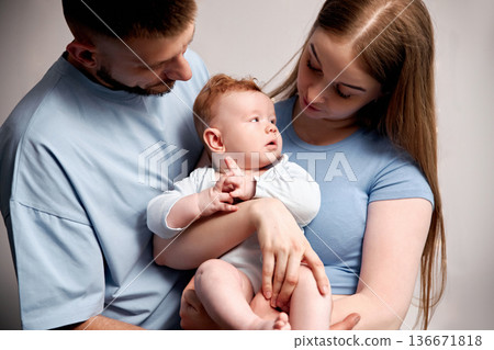 Family poses with baby in studio setting during daytime, showing warmth and connection in their interaction and expressions while holding the infant in their arms Family poses with baby in studio setting during daytime, showing warmth and connection in their interaction and expressions while holding the infant in their arms 136671818
