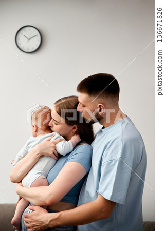 Family of three stands together in a cozy living room while holding their baby near a wall clock showing daytime hours 136671826