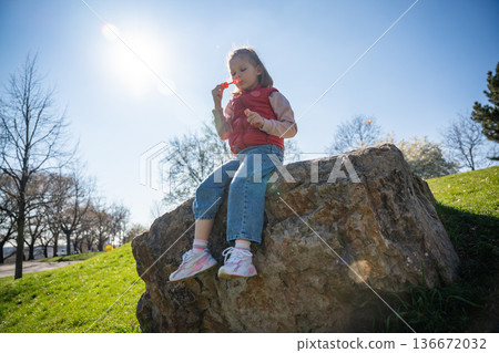 Little girl sitting on decorative stones blowing soap bubbles in city park. Spring childhood leisure and joyful outdoor play. Little girl sitting on decorative stones blowing soap bubbles in city park. Spring childhood leisure and joyful outdoor play. 136672032