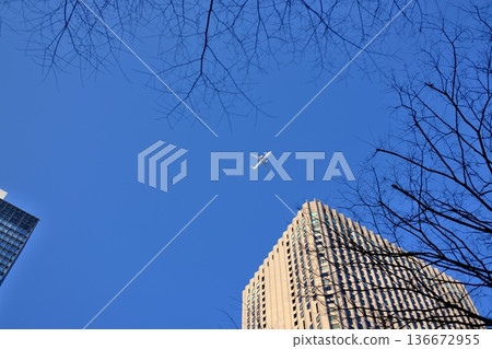 A white aircraft flying across the vast blue sky surrounded by city buildings A white aircraft flying across the vast blue sky surrounded by city buildings 136672955