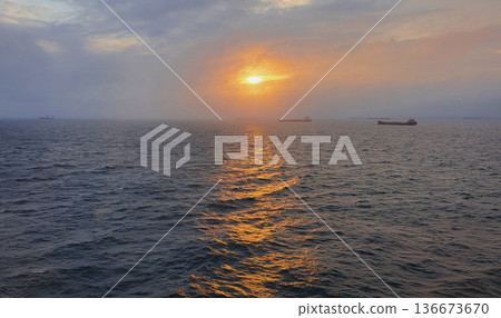 Dramatic pre-sunset seascape golden sunlight breaking through dense gray clouds, illuminating rippling dark blue water distant cargo ships on horizon 136673670