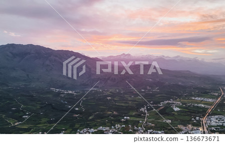 Aerial panoramic view Crete foothills at sunset with snow-capped peaks, olive groves, valley with lake scattered villages, winding road layered clouds Aerial panoramic view Crete foothills at sunset with snow-capped peaks, olive groves, valley with lake scattered villages, winding road layered clouds 136673671