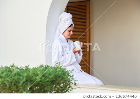 A woman in a robe and towel drinks a hot drink on a terrace after a shower A woman in a robe and towel drinks a hot drink on a terrace after a shower 136674440