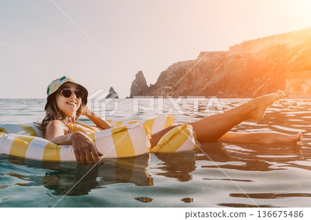 Woman Beach Float Relaxation - A woman wearing a bucket hat and sunglasses relaxes on a yellow and white striped inflatable float in the ocean. Woman Beach Float Relaxation - A woman wearing a bucket hat and sunglasses relaxes on a yellow and white striped inflatable float in the ocean. 136675486
