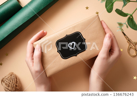 Hands holding a wrapped present with a heart on the label. Surrounded by gift wrapping supplies. 136676243