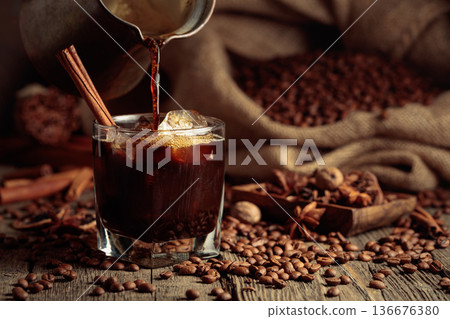 Black coffee is poured from a Turkish copper coffee pot into a glass with ice. 136676380