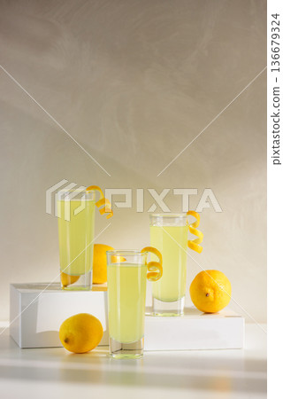 Limoncello Glasses with Curled Lemon Peels and Fresh Lemons on white platforms. Refreshing Summer Italian Beverage 136679324