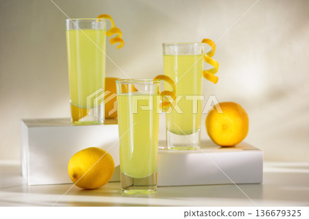 Limoncello Glasses with Curled Lemon Peels and Fresh Lemons on white platforms. Refreshing Summer Italian Beverage 136679325