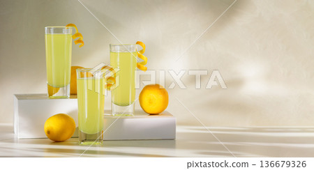 Limoncello Glasses with Curled Lemon Peels and Fresh Lemons on white platforms. Refreshing Summer Italian Beverage 136679326