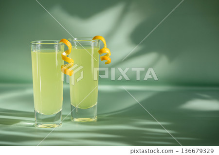 Limoncello Glasses with Curled Lemon Peels on Light Green Background. Refreshing Summer Italian Beverage 136679329