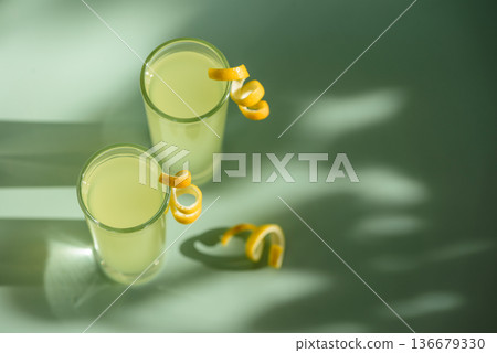 Limoncello Glasses with Curled Lemon Peels on Light Green Background. Refreshing Summer Italian Beverage 136679330