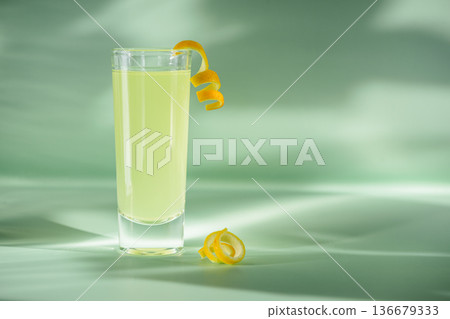 Limoncello Glasses with Curled Lemon Peels on Light Green Background. Refreshing Summer Italian Beverage 136679333