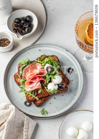 Roasted toasts with prosciutto ham, mozzarella balls, arugula and sliced olives on a plate on white table background. Delicious breakfast or snack 136679606