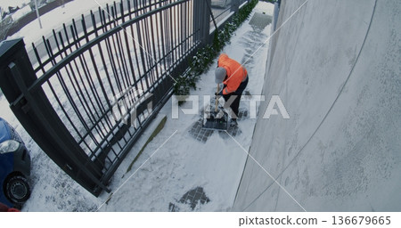 Male Utility Worker Wearing Reflective Jacket Removes Snow From Paved Walkway Beside Fence 136679665