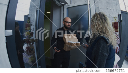 Female Courier With Headset Delivers Hot Coffee Cups and Brown Paper Bag to Man Doorstep 136679751