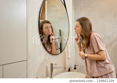 Woman standing at sink and examining face in bathroom mirror Woman standing at sink and examining face in bathroom mirror 136681104
