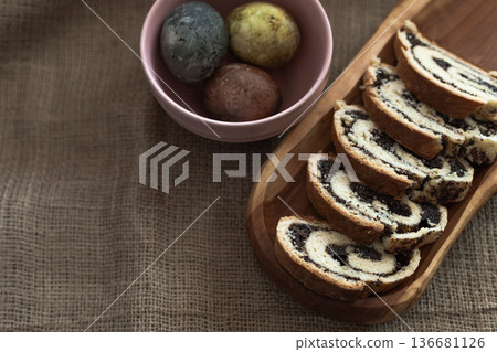 Traditional Easter eggs in a ceramic bowl and sliced poppy seed roll on a wooden plate over rustic burlap background 136681126