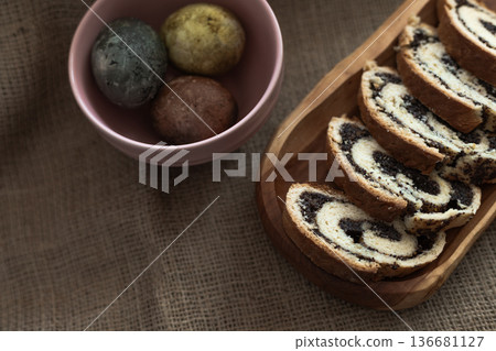 Colorful Easter eggs in a bowl beside sliced homemade poppy seed roulade on a wooden serving plate, rustic sackcloth backdrop Colorful Easter eggs in a bowl beside sliced homemade poppy seed roulade on a wooden serving plate, rustic sackcloth backdrop 136681127