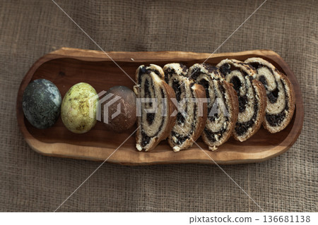 Sliced homemade poppy seed roll and three colorful Easter eggs arranged on a wooden plate, rustic holiday composition 136681138