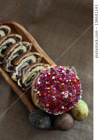 Overhead view of a festive Easter arrangement with three dyed eggs, homemade glazed Easter cake and neatly sliced poppy seed roll displayed on a long wooden serving board, rustic natural background Overhead view of a festive Easter arrangement with three dyed eggs, homemade glazed Easter cake and neatly sliced poppy seed roll displayed on a long wooden serving board, rustic natural background 136681141