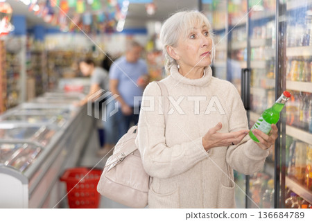 Old woman purchaser choosing cool drink in big supermarket Old woman purchaser choosing cool drink in big supermarket 136684789