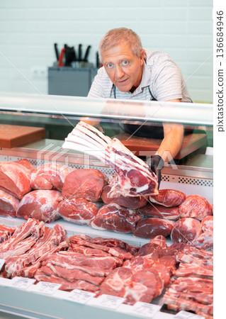 Positive elderly salesman in apron and disposable gloves shows raw beef tomahawk steak 136684946