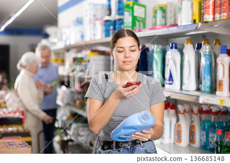 Young female customer scanning barcode on plastic bottle of household detergent with smartphone while shopping in supermarket, paying for item using mobile app 136685148